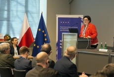 podsekretarz stanu w Ministerstwie Rodziny, Pracy i Polityki Społecznej Elżbieta Bojanowska przemawia z mównicy w auli KSAP. Widoczni siedzący uczestnicy konferencji. Obok mównicy baner KSAP i flagi polska i unijna.