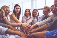 grupa uśmiechniętych kobiet stoi w kręgu i z wysuniętymi przed siebie rękoma, łączącymi się przed nimi